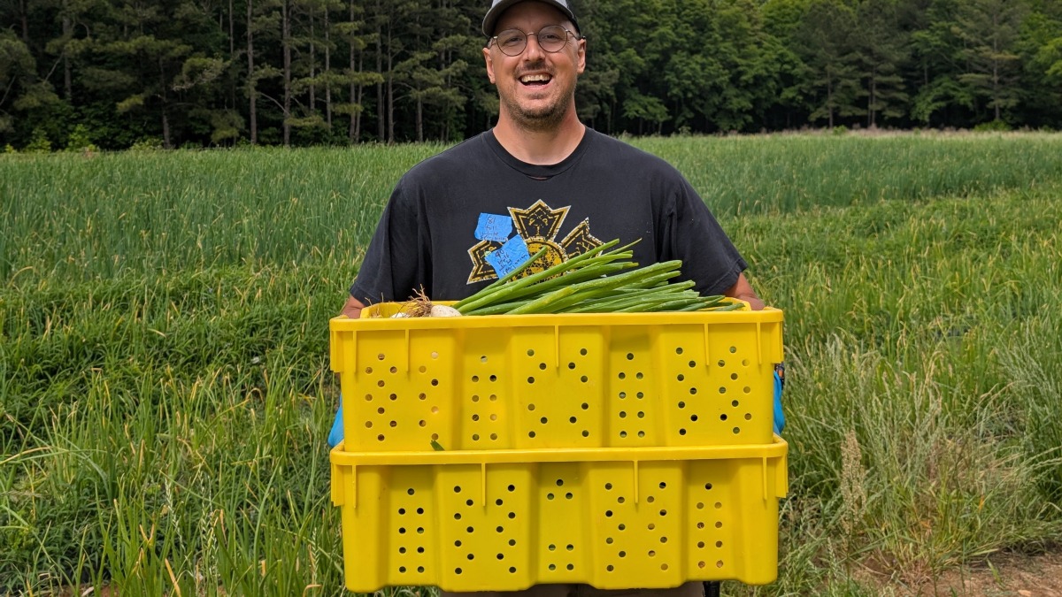 Smiling man standing in field holding stacked bins full of fresh produce.
