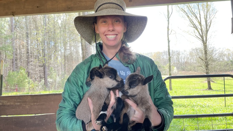 Woman wearing sunhat, green jacket, and holding 3 goat kids while smiling.